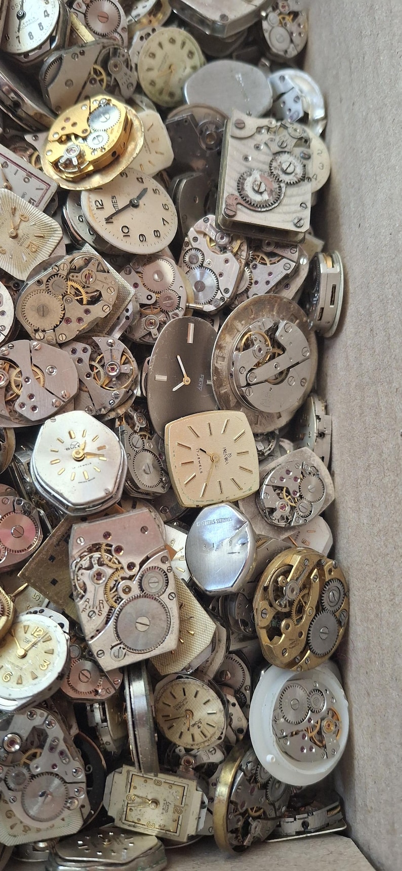 K&ouml;nnte beinhalten: Eine Sammlung von Vintage-Uhrenteilen, darunter Zifferbl&auml;tter, Zahnr&auml;der und Mechanismen. Die Zifferbl&auml;tter haben verschiedene Formen, darunter rund, quadratisch und rechteckig. Die Zahnr&auml;der sind aus Metall und haben komplizierte Designs. Die Teile befinden sich in einem Karton.
