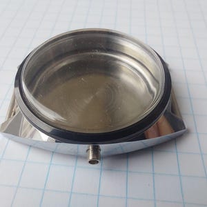 May include: A stainless steel watch case with a clear crystal and a black rubber gasket. The case has a rectangular shape with a circular opening for the watch face and a small tube extending from the bottom.