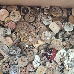 K&ouml;nnte beinhalten: Eine Kiste voller Vintage-Uhrenteile, darunter Zifferbl&auml;tter, Zahnr&auml;der und Mechanismen. Die Zifferbl&auml;tter zeigen verschiedene Stile und Farben, mit sichtbaren Zeigern und Ziffern. Die Teile weisen unterschiedliche Abnutzungserscheinungen auf.