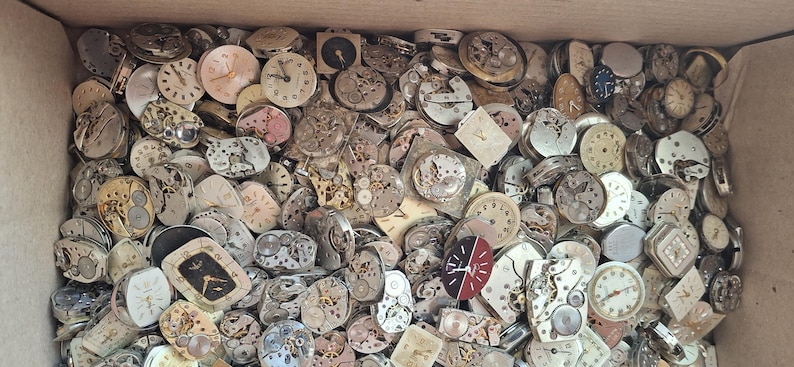 K&ouml;nnte beinhalten: Eine Kiste voller Vintage-Uhrenteile, darunter Zifferbl&auml;tter, Zahnr&auml;der und Mechanismen. Die Zifferbl&auml;tter zeigen verschiedene Stile und Farben, mit sichtbaren Zeigern und Ziffern. Die Teile weisen unterschiedliche Abnutzungserscheinungen auf.