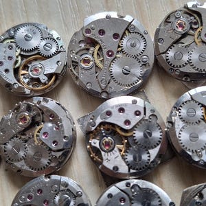 May include: A collection of disassembled watch movements, showcasing intricate gears, springs, and jewels. The silver-toned components are arranged in a grid pattern, revealing the inner workings of timepieces. The small, round mechanisms are displayed on a light wooden surface.