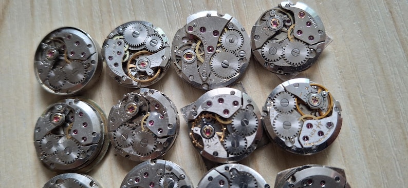 May include: A collection of disassembled watch movements, showcasing intricate gears, springs, and jewels. The silver-toned components are arranged in a grid pattern, revealing the inner workings of timepieces. The small, round mechanisms are displayed on a light wooden surface.