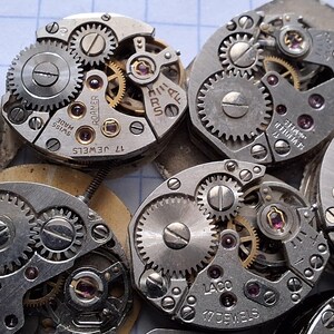May include: Close-up of several vintage watch movements, showcasing intricate gears, screws, and jewels. The silver-toned mechanisms are arranged in a cluster, highlighting the detailed craftsmanship of antique timepieces. Some movements have text like "17 Jewels" and "Laco".