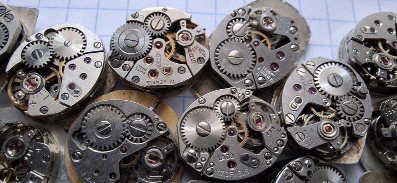 May include: Close-up of several vintage watch movements, showcasing intricate gears, screws, and jewels. The silver-toned mechanisms are arranged in a cluster, highlighting the detailed craftsmanship of antique timepieces. Some movements have text like "17 Jewels" and "Laco".