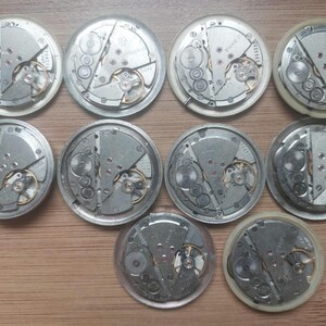 Pode incluir: Uma cole&ccedil;&atilde;o de dez movimentos de rel&oacute;gios vintage, mostrando engrenagens intrincadas e componentes mec&acirc;nicos. As pe&ccedil;as do rel&oacute;gio, em tons de prata, est&atilde;o dispostas sobre uma superf&iacute;cie de madeira, oferecendo um vislumbre do artesanato relojoeiro.