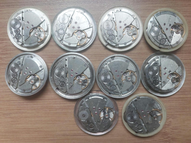 Pode incluir: Uma cole&ccedil;&atilde;o de dez movimentos de rel&oacute;gios vintage, mostrando engrenagens intrincadas e componentes mec&acirc;nicos. As pe&ccedil;as do rel&oacute;gio, em tons de prata, est&atilde;o dispostas sobre uma superf&iacute;cie de madeira, oferecendo um vislumbre do artesanato relojoeiro.