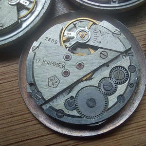 Pode incluir: Close-up de um movimento de rel&oacute;gio vintage com engrenagens, molas e um balan&ccedil;o intrincados. Os componentes met&aacute;licos s&atilde;o prateados e dourados, com o texto "2609" e texto cir&iacute;lico. O mecanismo do rel&oacute;gio est&aacute; sobre uma base circular.