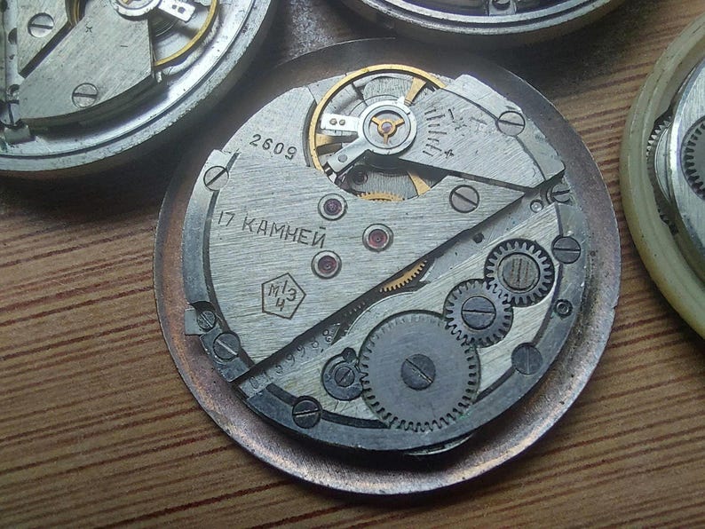 Pode incluir: Close-up de um movimento de rel&oacute;gio vintage com engrenagens, molas e um balan&ccedil;o intrincados. Os componentes met&aacute;licos s&atilde;o prateados e dourados, com o texto "2609" e texto cir&iacute;lico. O mecanismo do rel&oacute;gio est&aacute; sobre uma base circular.