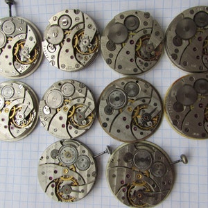 May include: A collection of 10 silver-toned watch movements with intricate gears and mechanisms. The movements are arranged in a grid pattern on a white gridded surface.