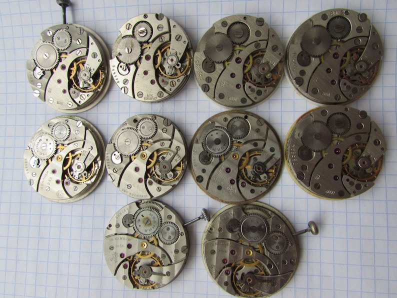 May include: A collection of 10 silver-toned watch movements with intricate gears and mechanisms. The movements are arranged in a grid pattern on a white gridded surface.