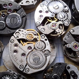 May include: Close-up of several vintage watch movements, showcasing intricate gears, jewels, and metal components. The watch parts are arranged in a scattered pattern, highlighting the mechanical details and craftsmanship of the timepieces.