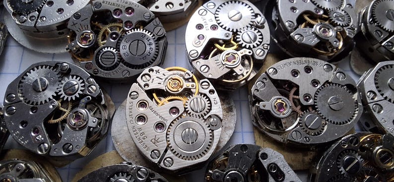 May include: Close-up of several vintage watch movements, showcasing intricate gears, jewels, and metal components. The watch parts are arranged in a scattered pattern, highlighting the mechanical details and craftsmanship of the timepieces.