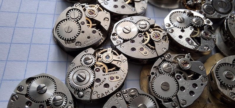 May include: Close-up of several disassembled watch movements, showcasing intricate gears, screws, and jewel bearings. The silver-toned metal components are arranged on a grid-lined surface, highlighting the precision of the watchmaking craft. Some movements are marked with "17 JEWELS" and "CAMY WATCH CO."
