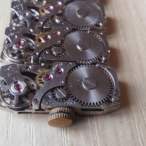 May include: Close-up of a watch movement, showcasing intricate gears, jewels, and metal components. The silver-toned parts are arranged in a rectangular pattern, with a small, gold-colored knob at the end. The background is a light-colored wooden surface.