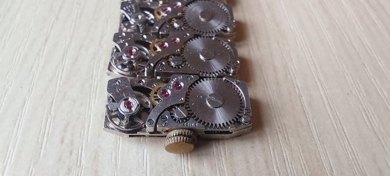 May include: Close-up of a watch movement, showcasing intricate gears, jewels, and metal components. The silver-toned parts are arranged in a rectangular pattern, with a small, gold-colored knob at the end. The background is a light-colored wooden surface.