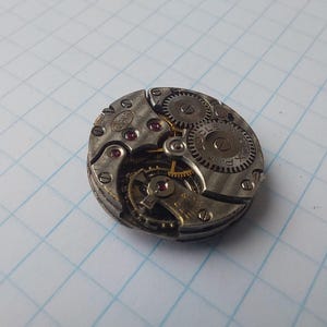 May include: Close-up of a vintage watch movement with visible gears, screws, and jewels. The silver-toned metal components are arranged in a circular pattern. The watch mechanism is set against a grid background, showcasing the intricate details of the timepiece.
