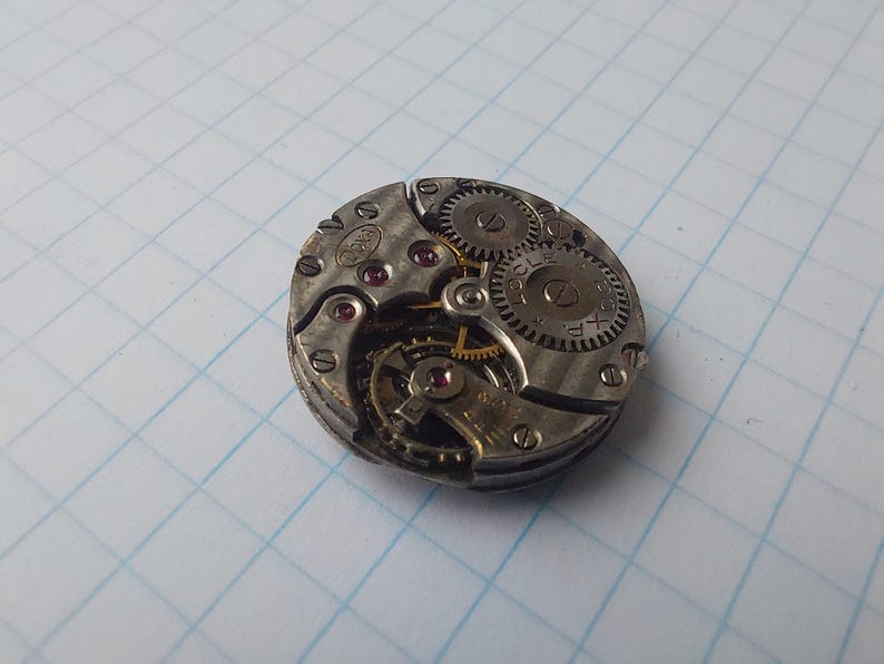 May include: Close-up of a vintage watch movement with visible gears, screws, and jewels. The silver-toned metal components are arranged in a circular pattern. The watch mechanism is set against a grid background, showcasing the intricate details of the timepiece.