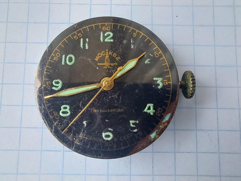 Puede incluir: Una esfera de reloj vintage con esfera negra, n&uacute;meros y manecillas verdes. La esfera del reloj tiene un marco dorado y una corona de cuerda en el lateral. El texto "MOCKBA" es visible en la parte superior.