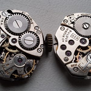 Puede incluir: Dos movimientos de reloj Bulova vintage, que muestran intrincados engranajes, joyas y el nombre de la marca. Los movimientos son de color plateado con componentes mecánicos visibles, incluyendo pequeños tornillos y dientes de engranaje. El texto "U.S.A." y "17 Jewels" son visibles.
