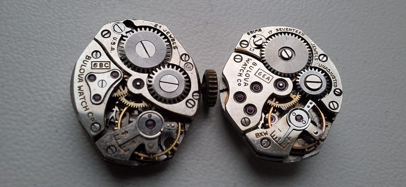 Peut inclure: Deux mouvements de montre Bulova vintage, pr&eacute;sentant des engrenages complexes, des bijoux et le nom de la marque. Les mouvements sont de couleur argent&eacute;e avec des composants m&eacute;caniques visibles, dont de petites vis et des dents d'engrenage. Le texte "U.S.A." et "17 Jewels" sont visibles.