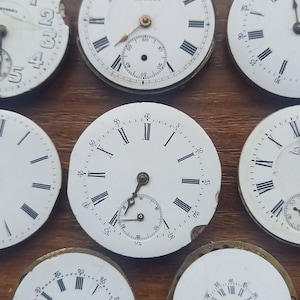 May include: A collection of antique watch faces with Roman numeral hour markers and sub-dials. The white faces have black markings and are set against a brown wooden surface. Some faces are damaged.