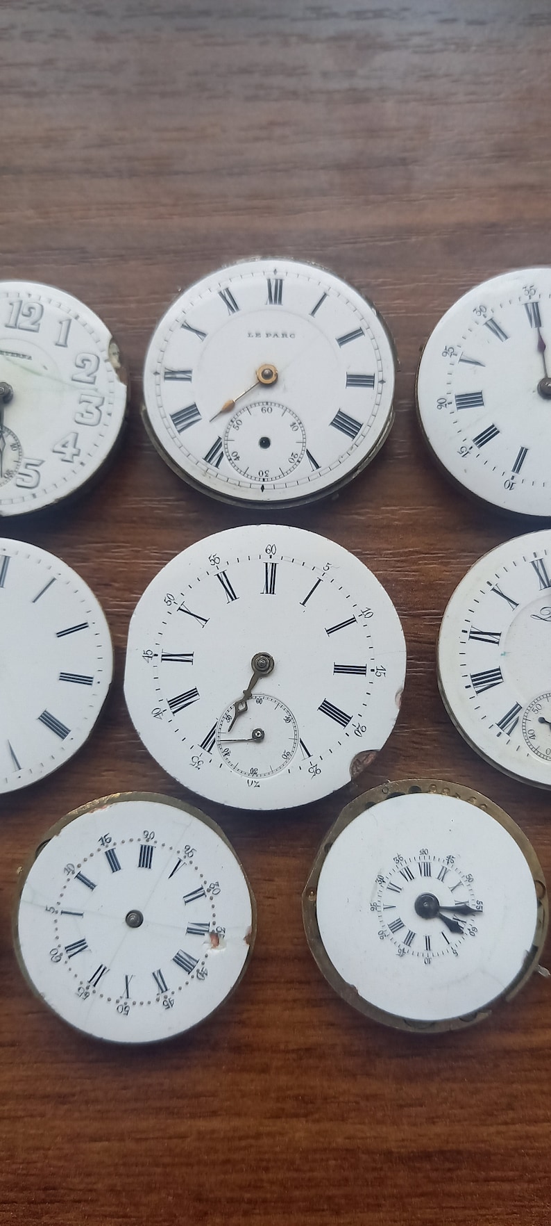 May include: A collection of antique watch faces with Roman numeral hour markers and sub-dials. The white faces have black markings and are set against a brown wooden surface. Some faces are damaged.