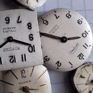 May include: A collection of vintage watch faces in various shapes, including square and round. The faces are silver-toned with black hands and numerals. Some faces have brand names like "Dugena" and "Anker" printed on them. The watch faces are scattered on a light blue grid.