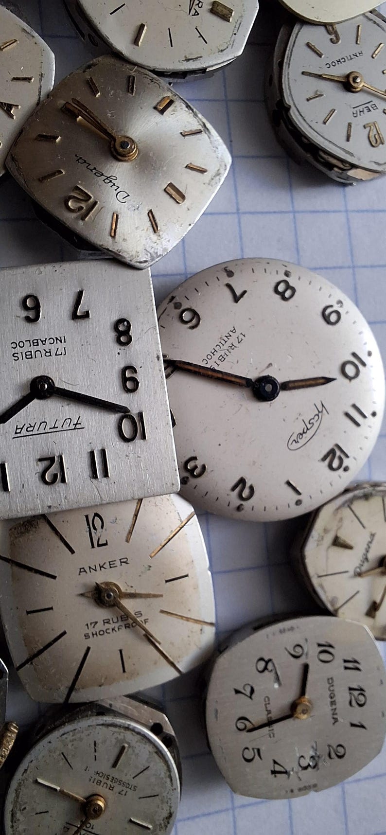 May include: A collection of vintage watch faces in various shapes, including square and round. The faces are silver-toned with black hands and numerals. Some faces have brand names like "Dugena" and "Anker" printed on them. The watch faces are scattered on a light blue grid.