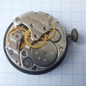 Puede incluir: Primer plano de un mecanismo de reloj con engranajes, tornillos y una tija visibles. Los componentes met&aacute;licos son plateados y dorados, con el texto "6 камней". El mecanismo del reloj est&aacute; sobre un fondo cuadriculado.