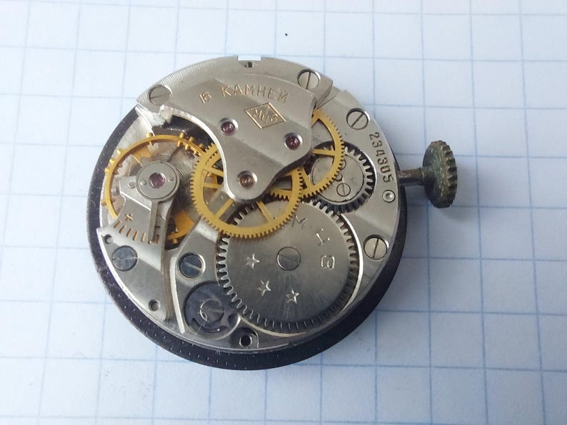 Puede incluir: Primer plano de un mecanismo de reloj con engranajes, tornillos y una tija visibles. Los componentes met&aacute;licos son plateados y dorados, con el texto "6 камней". El mecanismo del reloj est&aacute; sobre un fondo cuadriculado.