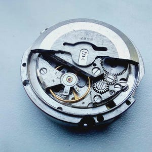 Op de afbeelding: Een rond mechanisch horloge-uurwerk met een zilverkleurige oscillerende massa met het gegraveerde nummer 2427. Het interne mechanisme toont diverse metalen tandwielen, een goudkleurig balanswiel en kleine schroeven in een metalen frame.