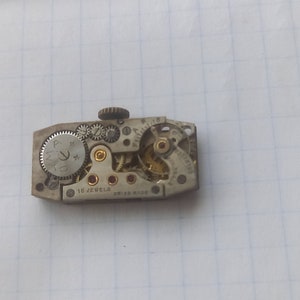 May include: Close-up of a silver watch movement with 15 jewels and Swiss Made markings. The movement is exposed, showing the gears and other internal components.