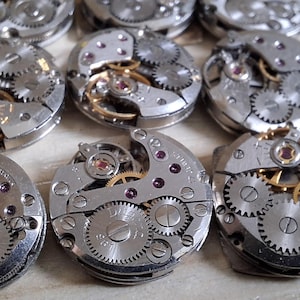 May include: A collection of disassembled watch movements, showcasing intricate gears, jewels, and metallic components. The silver-toned mechanisms are arranged in a close-up shot, highlighting the precision engineering of the watch parts.
