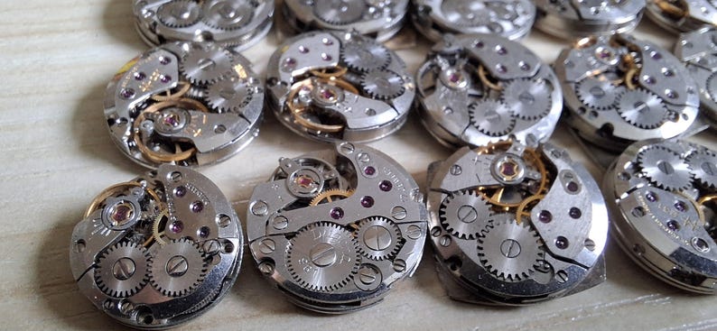 May include: A collection of disassembled watch movements, showcasing intricate gears, jewels, and metallic components. The silver-toned mechanisms are arranged in a close-up shot, highlighting the precision engineering of the watch parts.