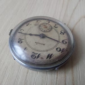 Puede incluir: Reloj de pulsera Zenith vintage con caja plateada y esfera crema. El reloj presenta n&uacute;meros negros, manecillas y el logotipo de Zenith. El reloj redondo tiene una cubierta de cristal transparente.