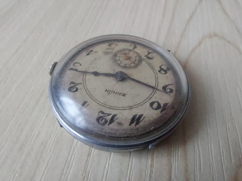 Puede incluir: Reloj de pulsera Zenith vintage con caja plateada y esfera crema. El reloj presenta n&uacute;meros negros, manecillas y el logotipo de Zenith. El reloj redondo tiene una cubierta de cristal transparente.