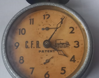 May include: Vintage alarm clock with a round face, featuring a cream-colored dial with black numerals and hands. The clock has a metal casing, a bell on top, and the text "C.F.R. PATENT".