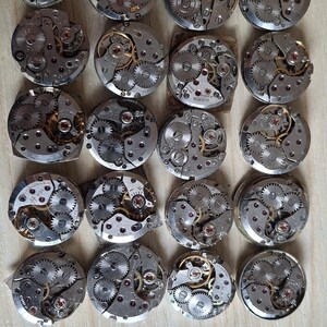 May include: A collection of vintage watch movements, showcasing intricate gears, jewels, and metal components. The circular mechanisms are arranged on a light wooden surface, highlighting the detailed craftsmanship of each piece.