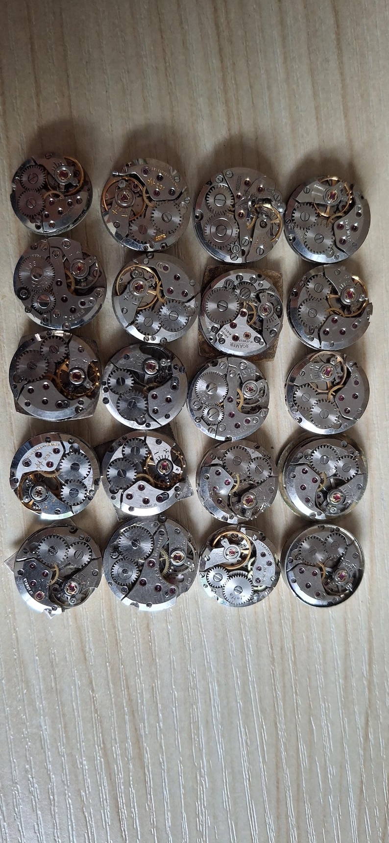 May include: A collection of vintage watch movements, showcasing intricate gears, jewels, and metal components. The circular mechanisms are arranged on a light wooden surface, highlighting the detailed craftsmanship of each piece.