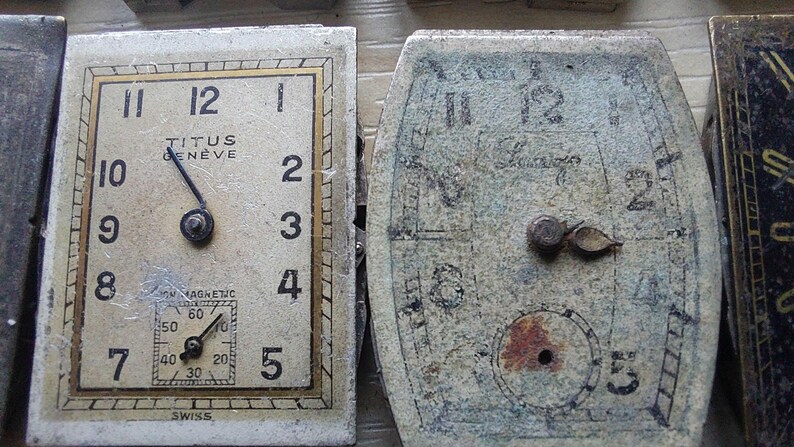 Peut inclure: Deux cadrans de montre vintage. Le cadran de gauche est rectangulaire avec le texte "TITUS GENEVE" et le mot "SWISS". Le cadran de droite est ovale et tr&egrave;s us&eacute;. Les deux cadrans affichent des chiffres et des aiguilles.
