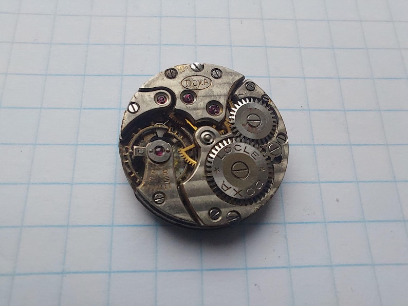 May include: Close-up of a vintage watch movement with intricate gears, screws, and ruby jewels. The watch mechanism is circular, with the brand name "DOXA" visible. The gears are silver-toned, and the jewels are red.