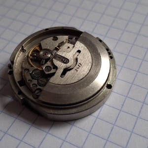 K&ouml;nnte beinhalten: Nahaufnahme eines zerlegten Uhrwerks, das Zahnr&auml;der, Federn und die Zahl "2427" auf einem Metallbauteil zeigt. Der silberfarbene Uhrenmechanismus ist vor einem karierten Hintergrund zu sehen.