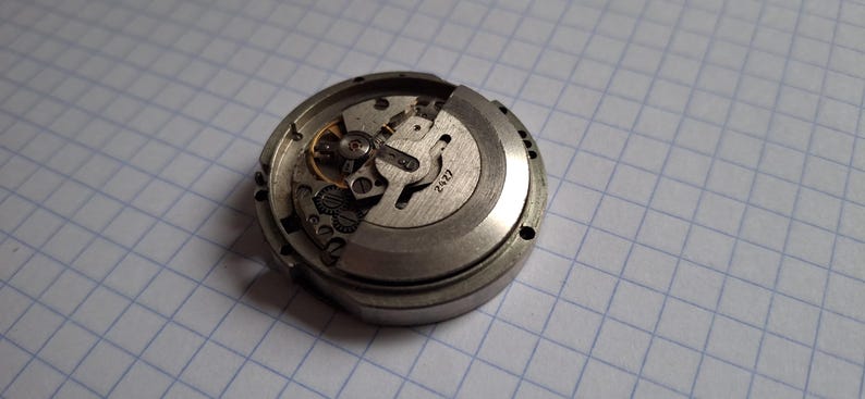 K&ouml;nnte beinhalten: Nahaufnahme eines zerlegten Uhrwerks, das Zahnr&auml;der, Federn und die Zahl "2427" auf einem Metallbauteil zeigt. Der silberfarbene Uhrenmechanismus ist vor einem karierten Hintergrund zu sehen.