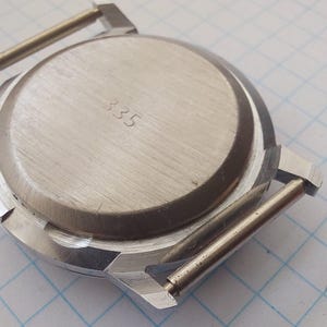 May include: A close-up of the back of a silver watch case. The case has a circular brushed metal back with the number "335" etched into it. The watch case has two silver bars for the watch strap and is set against a white and blue grid background.