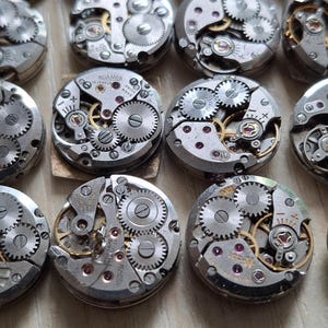 May include: A collection of vintage watch movements, showcasing intricate gears, springs, and jewels. The silver-toned mechanisms are arranged in a grid pattern, with visible details of the internal components. The watch parts are on a light brown surface.