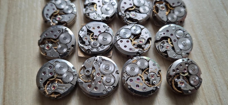 May include: A collection of vintage watch movements, showcasing intricate gears, springs, and jewels. The silver-toned mechanisms are arranged in a grid pattern, with visible details of the internal components. The watch parts are on a light brown surface.