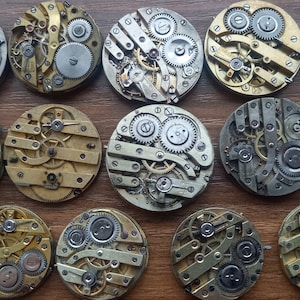 May include: A collection of vintage watch movements, showcasing intricate gears and mechanisms. The circular components are in various metallic tones, including silver, gold, and brass. The arrangement is on a wooden surface, highlighting the detailed craftsmanship.