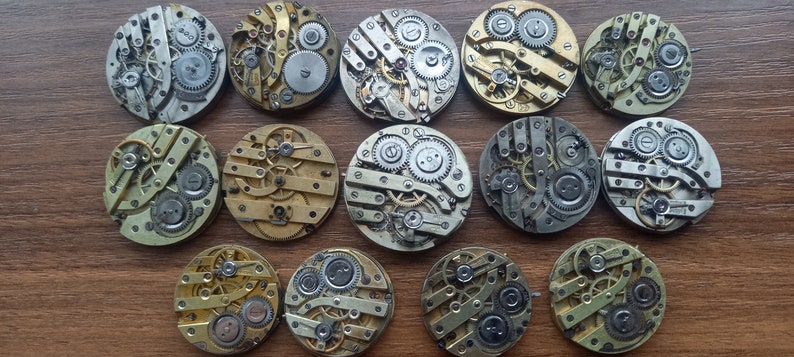 May include: A collection of vintage watch movements, showcasing intricate gears and mechanisms. The circular components are in various metallic tones, including silver, gold, and brass. The arrangement is on a wooden surface, highlighting the detailed craftsmanship.