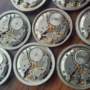 May include: Close-up of eight silver watch movements with intricate gears and mechanisms. The movements are arranged in a circular pattern on a wooden surface.