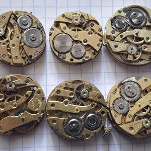 Op de afbeelding: Een verzameling van tien vintage uurwerken, met ingewikkelde messing tandwielen, veren en balanswielen. De cirkelvormige mechanismen zijn in twee rijen gerangschikt en tonen de complexe interne werking van antieke uurwerken.